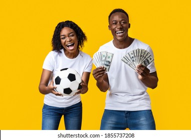 Sport Bet Excitement, Gambing And Family Lifestyle Concept. Excited African-american Young Couple Enjoying Their Win, Standing With Soccer Ball And Lots Of Money, Yellow Studio Background