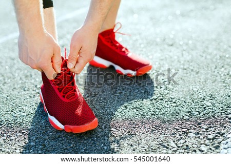 Sport concept, red sneakers for running. Man tightening lacings on his sneakers, no face. Legs in professional sport shoes, closeup. Outdoors, sunlight, stadium