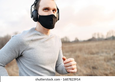 Sport during quarantine, self-isolation in the countryside. A young athletic guy is jogging on a dirt road in the meadow. He is wearing a black medical mask and headphones - Powered by Shutterstock