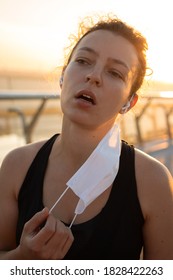 Sport During Quarantine,coronavirus, Covid-19 Concept. Tired Young Athletic Woman Runner Taking Off Medical Protective Mask And Free Breathing Deep, Taking A Rest After Workout Hard Outdoors.Vertical