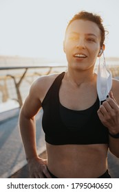 Sport During Quarantine,coronavirus. Smiling Tired Young Athletic Woman Runner Taking Off Medical Protective Mask And Free Breathing Deep. Taking A Rest After Running Workout Hard Outdoors. Vertical