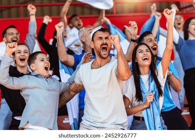 Sport fans cheering at the game on stadium. Wearing blue and white colors to support their team. Celebrating with flags and scarfs.