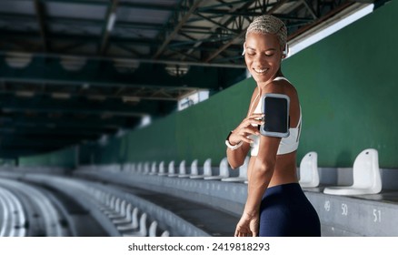 sport female athlete during running training and jogging, smiling African American woman touch a smartphone to armband, wear earphones and fitness smartwatch. Utilizing empty stadium stands and stairs - Powered by Shutterstock