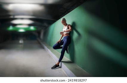 sport female athlete running training and jogging, African American woman look fitness smartwatch, wears earphones and armband. Leaning against green urban wall of hallway underground with copy space - Powered by Shutterstock