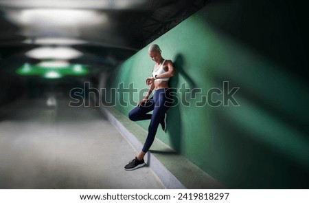 sport female athlete running training and jogging, African American woman look fitness smartwatch, wears earphones and armband. Leaning against green urban wall of hallway underground with copy space