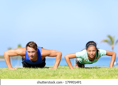 Sport Fitness Couple Doing Push Ups Outdoor. Young Athletes Doing Pushups Exercise Outside On Grass In Summer. Caucasian Man Sports Model And Asian Woman Fitness Model Doing Push-ups Workout.