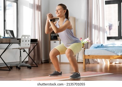 Sport, Fitness And Healthy Lifestyle Concept - Smiling Teenage Girl With Smart Watch Exercising On Yoga Mat And Doing Squats At Home