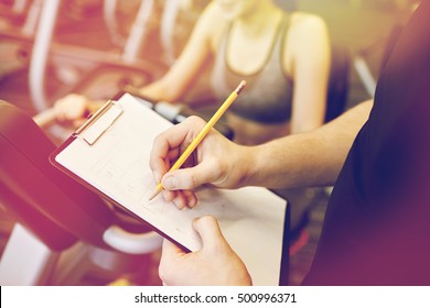 sport, fitness, lifestyle, technology and people concept - close up of trainer hands with clipboard writing and woman working out on exercise bike in gym - Powered by Shutterstock