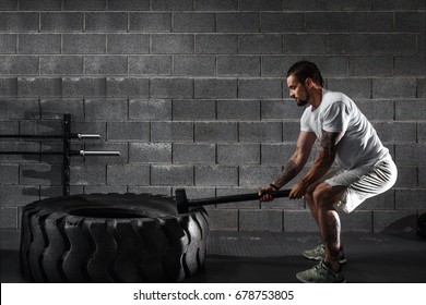 Sport Fitness Man Hitting Wheel Tire With Hammer Sledge Crossfit Training, Young Healthy Guy Gym Interior