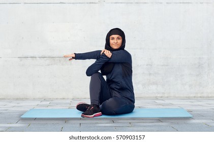 sport, fitness and people concept - happy muslim woman in hijab sitting and stretching on mat over gray concrete wall background - Powered by Shutterstock