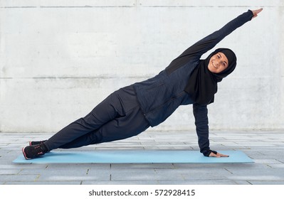 Sport, Fitness And People Concept - Happy Smiling Muslim Woman In Hijab Doing Plank Exercise On Mat Over Gray Concrete Wall Background