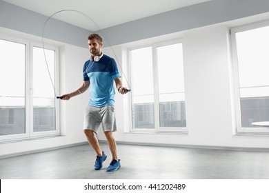 Sport And Fitness Workout. Healthy Athletic Man With Muscular Body In Fashion Headphones, Sportswear Skipping With Jump Rope, Exercising Indoor. Handsome Male Doing Jumping Cardio Exercise Training.