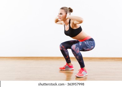 Sport girl with dark hair wearing pink snickers, dark leggings and black short top doing squatting at gym, fitness, white wall and wooden floor, copy space.