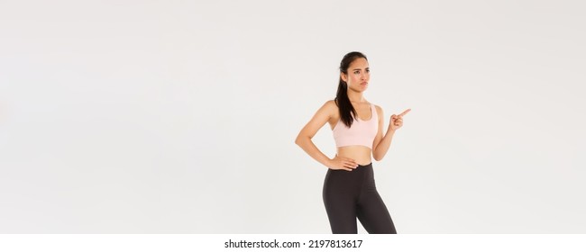 Sport, Gym And Healthy Body Concept. Full Length Of Skeptical And Displeased Female Athlete, Asian Sportswoman Sulking And Frowning Disappointed, Pointing Finger Upper Left Corner And Complaining