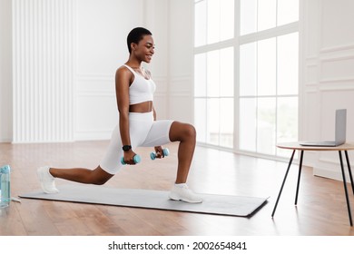 Sport And Healthy Lifestyle. Fit Black Woman Watching Training Tutorial On Laptop, Doing Forward Walking Lunges With Dumbbells, Exercising At Home. Smiling Lady Looking At Pc Screen On Tea Table