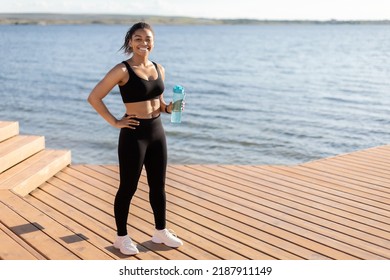 Sport And Hydration Concept. Happy Millennial Black Woman Training Next To Lake, Drinking Water While Having Break, Looking At Camera And Smiling, Panorama, With Copy Space, Full Length Shot