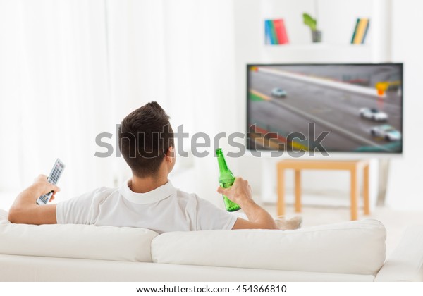 sport, leisure, technology, motorsports and people\
concept - man watching car racing on tv and drinking beer from\
bottle at home from back