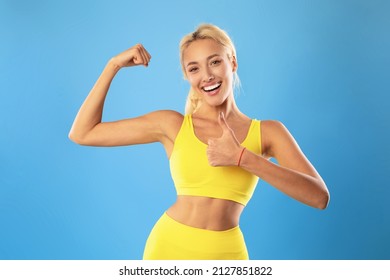 Sport Is Life. Portrait Of Excited Slim Woman In Yellow Sportswear Posing, Showing Biceps And Thumbs Up Sign Gesture After Good Workout Isolated On Blue Studio Background, Promoting Healthy Lifestyle