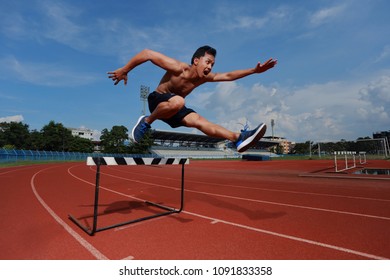 The Sport Man To Runner Jumping Over An Hurdle During Track And Field Event. Athlete Running A Hurdle Race In A Stadium.Dramatic Image