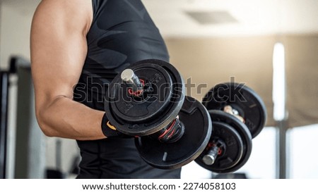 Sport man with well trained body in black sportswear wearing sport gloves lifting two dumbbells in fitness gym. Weight training and bodybuilding workout concept.