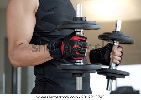 Sport man with well trained body in black sportswear wearing sport gloves lifting two dumbbells in fitness gym. Weight training and bodybuilding workout concept