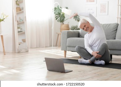 Sport In Mature Age. Happy senior woman doing stretching exercises in front of laptop at home, watching online tutorials, free space
