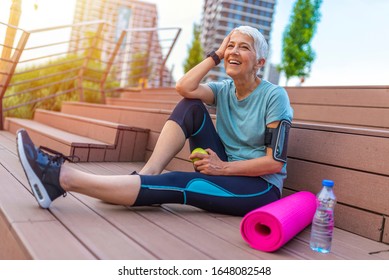 Sport Mature Woman Sitting And Resting After Workout Or Exercise And Eating Apple On Floor. Relax Concept. Strength Training And Body Build Up Theme. Beautiful Sporty Woman Eating Apple