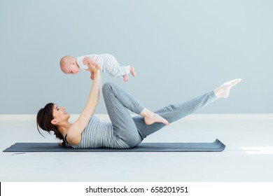 Sport, motherhood and active lifestyle concept - side view of young mother workout together with her baby over gray wall background. Mother having fun and playing with her little son. - Powered by Shutterstock