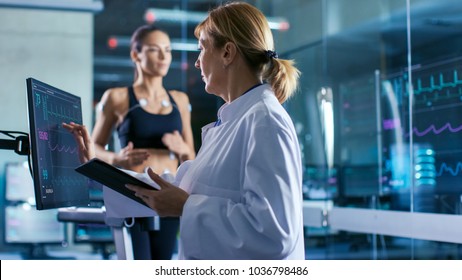 Sport Scientist Supervises Touches Display Showing EKG Status While in the Background Woman Athlete Running on a Treadmill with Electrodes Attached to His Body. Laboratory with High-Tech Equipment.