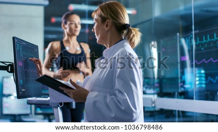 Sport Scientist Supervises Touches Display Showing EKG Status While in the Background Woman Athlete Running on a Treadmill with Electrodes Attached to His Body. Laboratory with High-Tech Equipment.