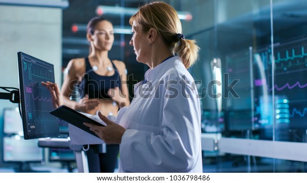 Sport Scientist Supervises Touches Display\
Showing EKG Status While in the Background Woman Athlete Running on\
a Treadmill with Electrodes Attached to His Body. Laboratory with\
High-Tech Equipment.