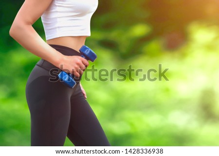 Sport training concept. Girl in white t-shirt holding a dumbbell in her hand on the background of the park. The girl is engaged in fitness in nature