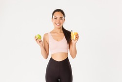 Sport, Wellbeing And Active Lifestyle Concept. Portrait Of Healthy And Fit Good-looking Asian Sportswoman Holding Orange Juice And Apple, Morning Breakfast Before Workout, White Background