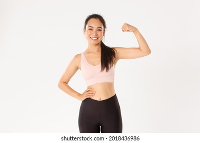 Sport, Wellbeing And Active Lifestyle Concept. Portrait Of Smiling Slim And Strong Asian Fitness Girl, Personal Workout Trainer Showing Muscles, Flexing Biceps And Look Proud, White Background