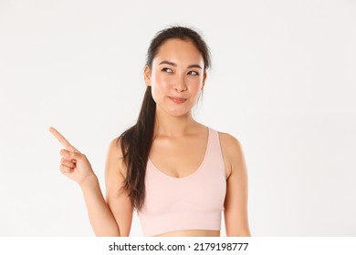 Sport, wellbeing and active lifestyle concept. Cunning and thoughtful smiling asian sportswoman, fitness girl making her choice, looking pleased with intrigued smile, pointing upper left corner