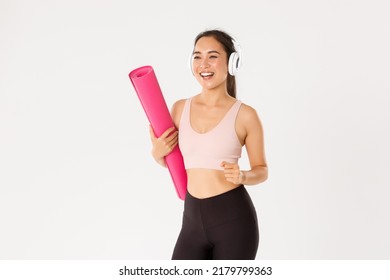 Sport, wellbeing and active lifestyle concept. Cheerful smiling asian fitness girl in headphones and sportswear going to gym with rubbermat, hurry up for yoga classes excited, white background - Powered by Shutterstock