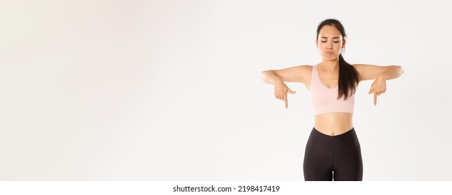 Sport, Wellbeing And Active Lifestyle Concept. Sulking And Disappointed Asian Workout Girl, Female Athlete In Activewear Frowning Upset, Pointing And Looking Down At Bad Thing, White Background