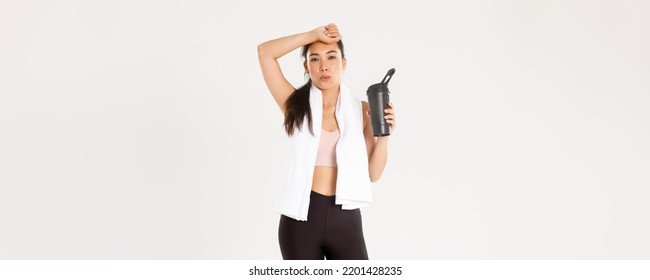 Sport, Wellbeing And Active Lifestyle Concept. Tired But Satisfied Asian Fitness Girl Wiping Sweat From Forehead And Exhale After Good Workout, Drinking Water Or Protein During Training Session