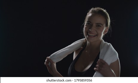 Sport Woman With Towel On Shoulders Posing On Black Background. Portrait Of Fitness Model With Towel Posing In Dark Gym. Close Up Of Happy Woman Smiling After Sport Workout. Sport People Concept