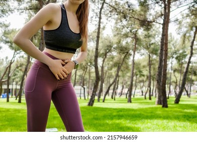 Sport woman wearing sports bra and yoga pants stomachache in a city park, outdoors in summer. Beautiful young woman have stomach ache. Outdoor sports and pain concepts.