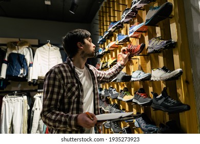 Sporting goods store owner with clipboard checking inventory. Tourist store manager working near showcase with hiking boots makes check list of orders. Salesman exhibit display of walking boots.
