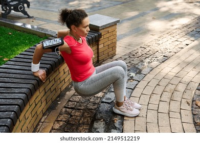 Sportive active middle-aged Hispanic woman exercising outdoor. Active sportswoman doing triceps exercises leaning her hands on a wooden bench in a city park