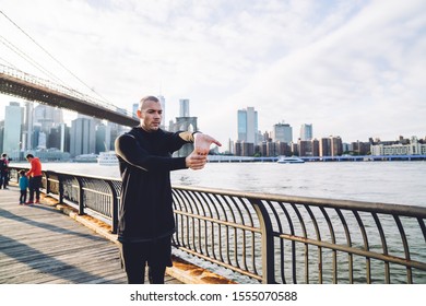 Sportive Adult Man In Black Sportswear Doing Wrist Stretch On Boardwalk Of City Embankment Against Modern Skyscrapers In Morning Sunlight 