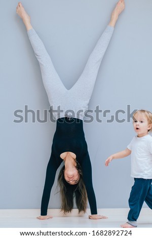 Sportive brunette fit mother playing with little baby son doing a handstand exercise at empty home or Lounge exercise. Conception of motherhood youth and energy. Vertical shoy. Copy space