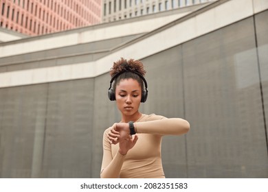 Sportive curly haired young woman stands outdoors rests after workout checks results on smartwatch listens music in headphones wears active wear leads sporty lifestyle. Fitness devices concept - Powered by Shutterstock