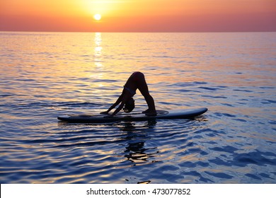 Sportive female doing yoga exercise on the beach over beautiful sunset background, standing on the sup board on the water in Pose dog muzzle down, Adho Mukha Svanasana