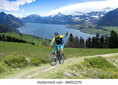 Sportive Man In Middle Age With Mountain Bike On Mountain Trail Beckons The Viewer. The Background Shows The Lake Lucerne.