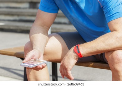 Sportive Man Sits On A Bench And Checks His Fitness Results On A Smart Phone. He Wears A Fitness Tracker Wristband On His Left Arm.