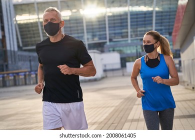 Sportive Middle Aged Couple Jogging With Protective Masks In The Morning. Covid19 Responsible Behavior