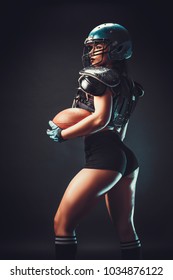 Sportive serious woman in helmet of rugby player holding ball in stuio on dark background.
